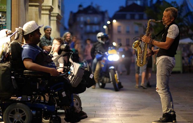 Mann im Rollstuhl hört einem Strassenmusikanten mit Saxophon zu.
